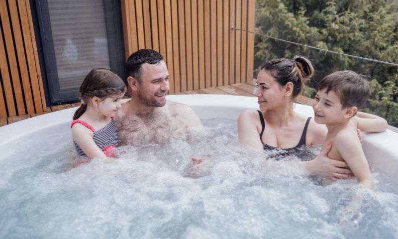 Happy family with two small children have fun in the jacuzzi. Cheerful young dad splashes water on his laughing cute son. Smiling man, woman, funny son and cute daughter relaxing in bubbling water
